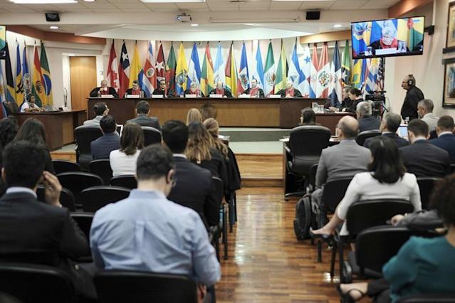 Imagen de personas en una corte, con el tribunal, el  publico sentado y banderas al fondo